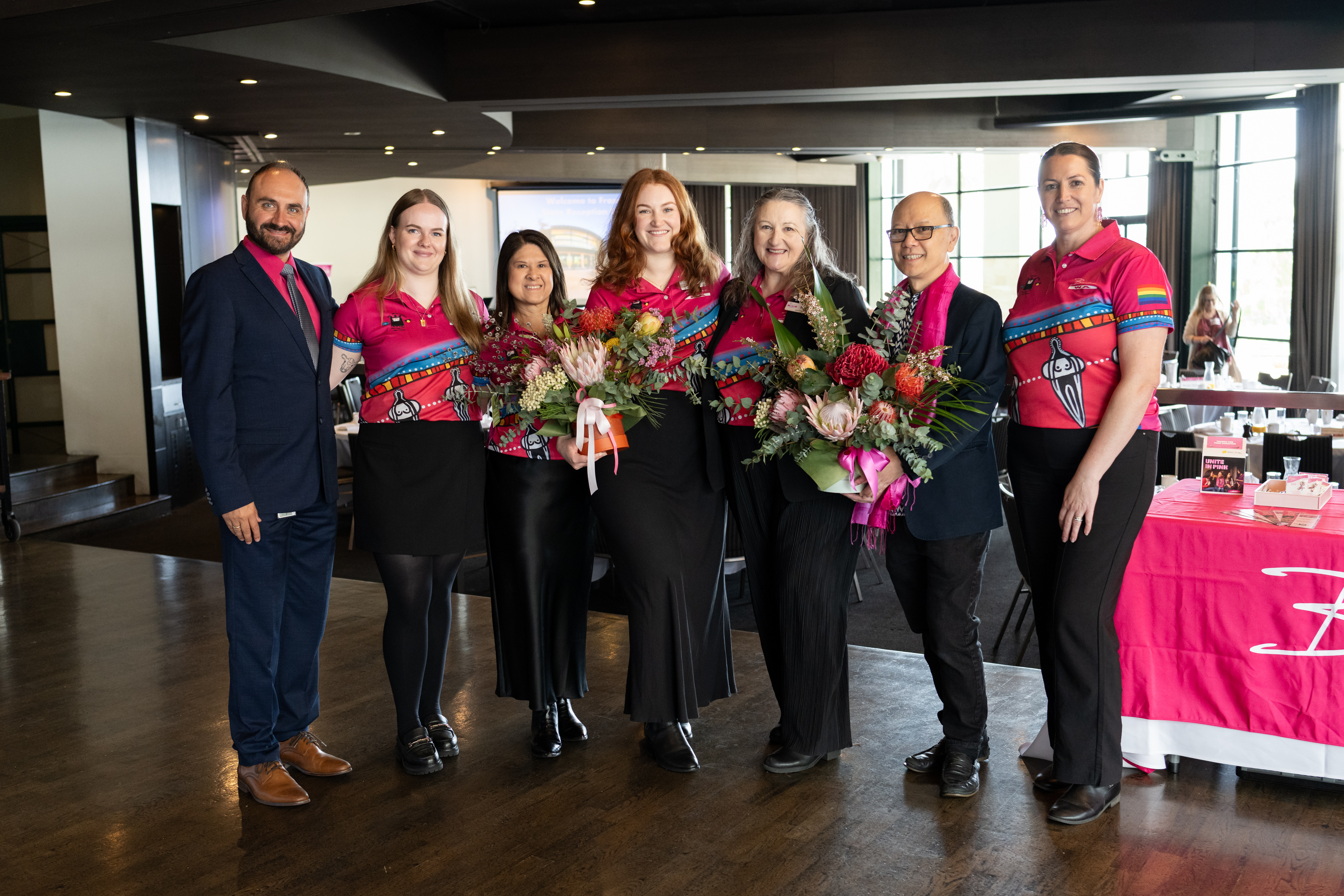 Health Promotion team standing in a line all with matching pink shirts and holding bouquet of flowers
