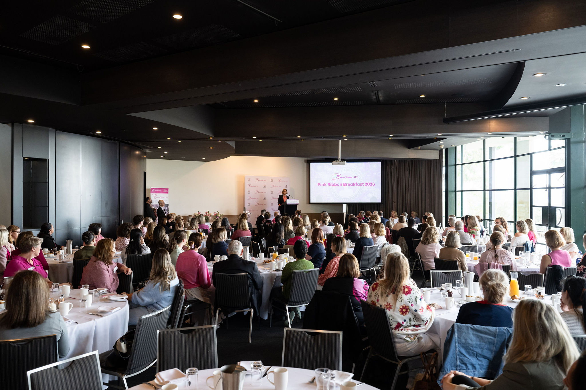 View from back of the room at Pink Ribbon Breakfast. Guests are seated at round tables facing the front of the room.