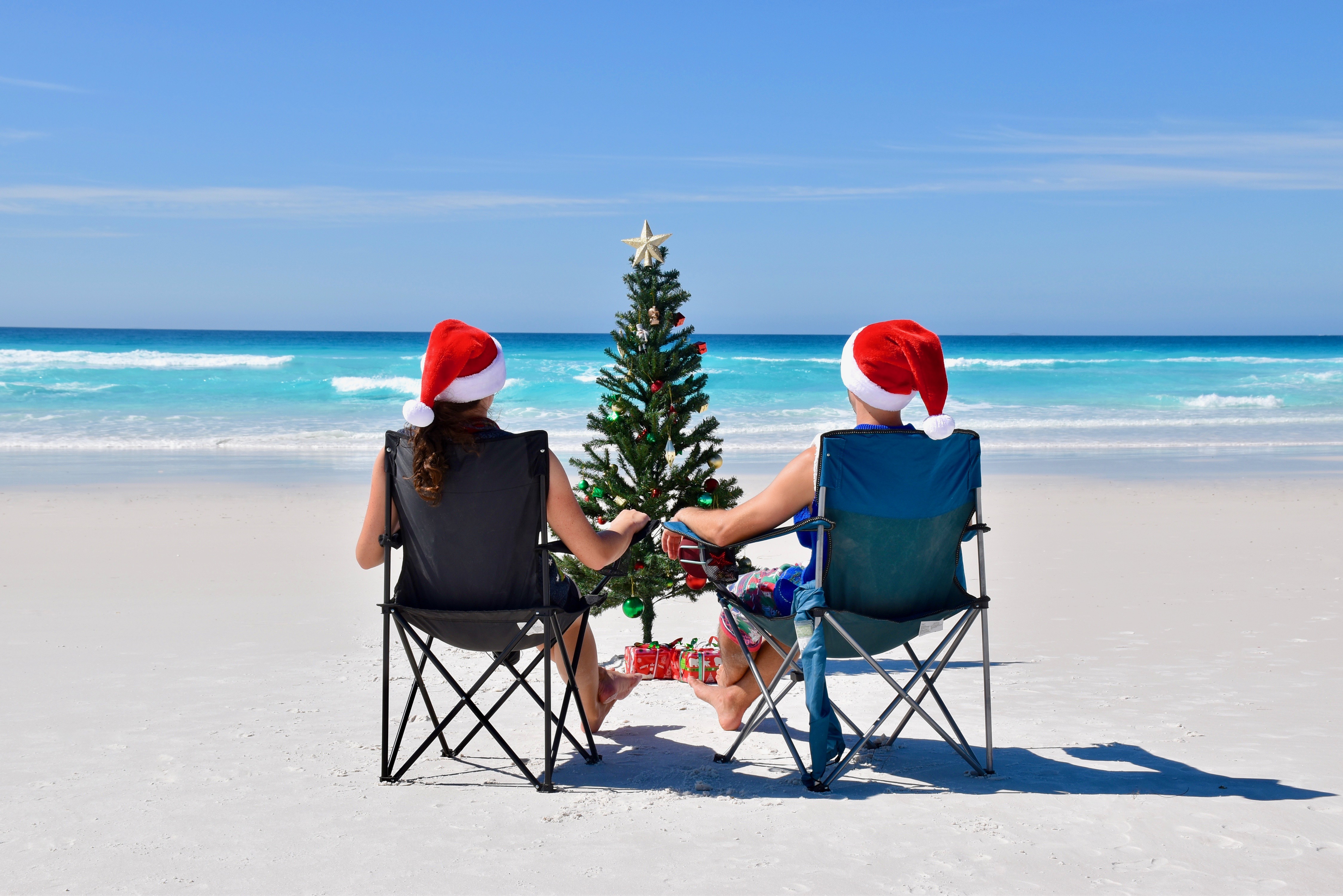 Two people at a sunny beach on camp chairs with a Christmas tree and Christmas hats