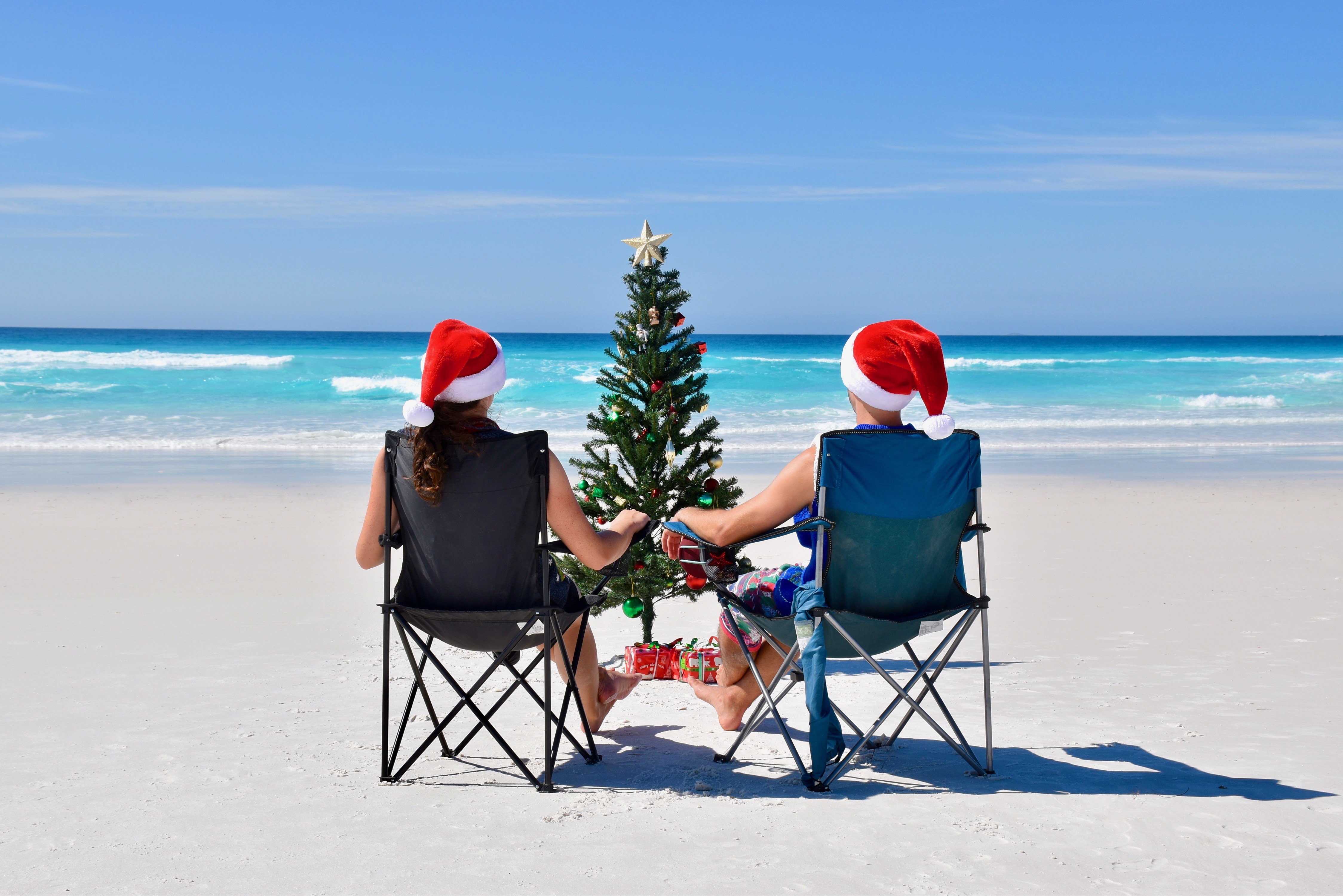 Two people at a sunny beach on camp chairs with a Christmas tree and Christmas hats